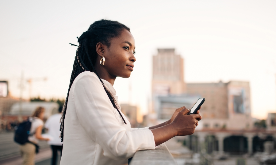 Mulher segura um celular ao entardecer, com a cidade ao fundo iluminada por tons dourados.