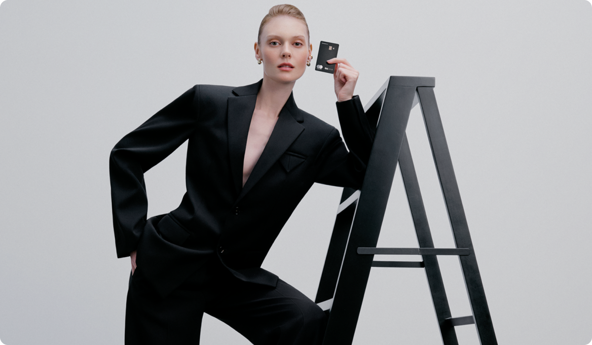 Mulher de terno preto posa com um Cartão C6 Carbon Mastercard Black, apoiada em uma escada.