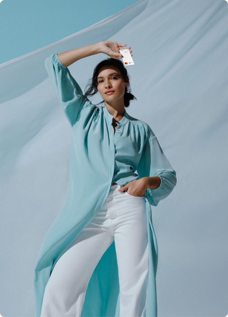 Mulher com blusa azul clara segura um Cartão C6 Bank, com fundo esvoaçante ao vento.