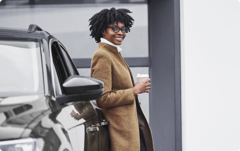Mulher sorridente de óculos, com casaco marrom, encostada em um carro e segurando um café.