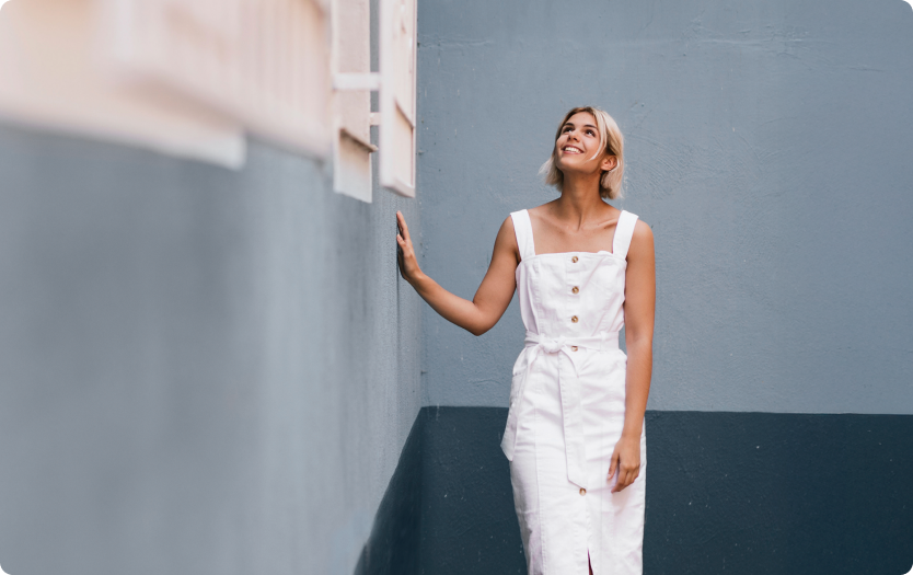 Mulher de vestido branco em corredor azul olha para cima com a mão apoiada na parede.