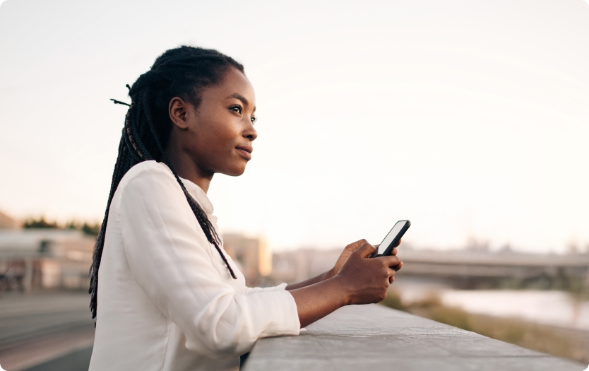 Mulher segura um celular e observa a cidade ao fundo.