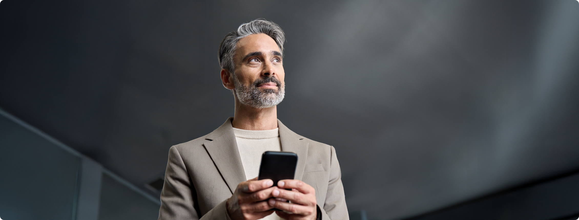 Homem de blazer claro olha para cima com celular nas mãos.