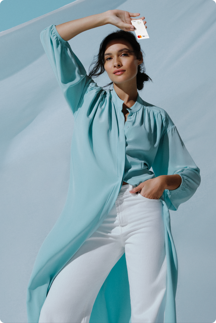 Foto de mulher branca, de cabelos castanhos, vestindo uma camisa azul clara e uma calça branca. Ela segura um cartão da C6 Conta Global branco acima da cabeça e expressa um leve sorriso. Atrás dela, um céu limpo e um tecido esvoaçante azul claro.