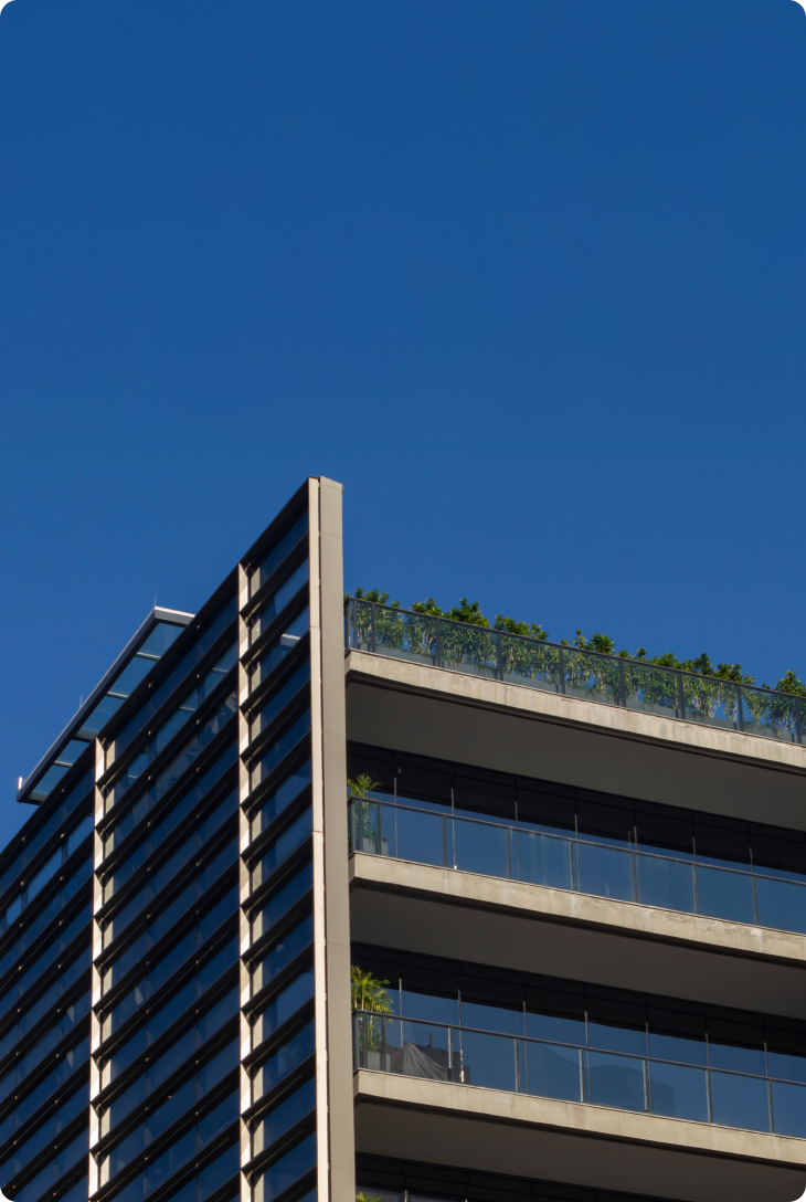 Prédio moderno com sacadas e plantas no topo, céu azul ao fundo.