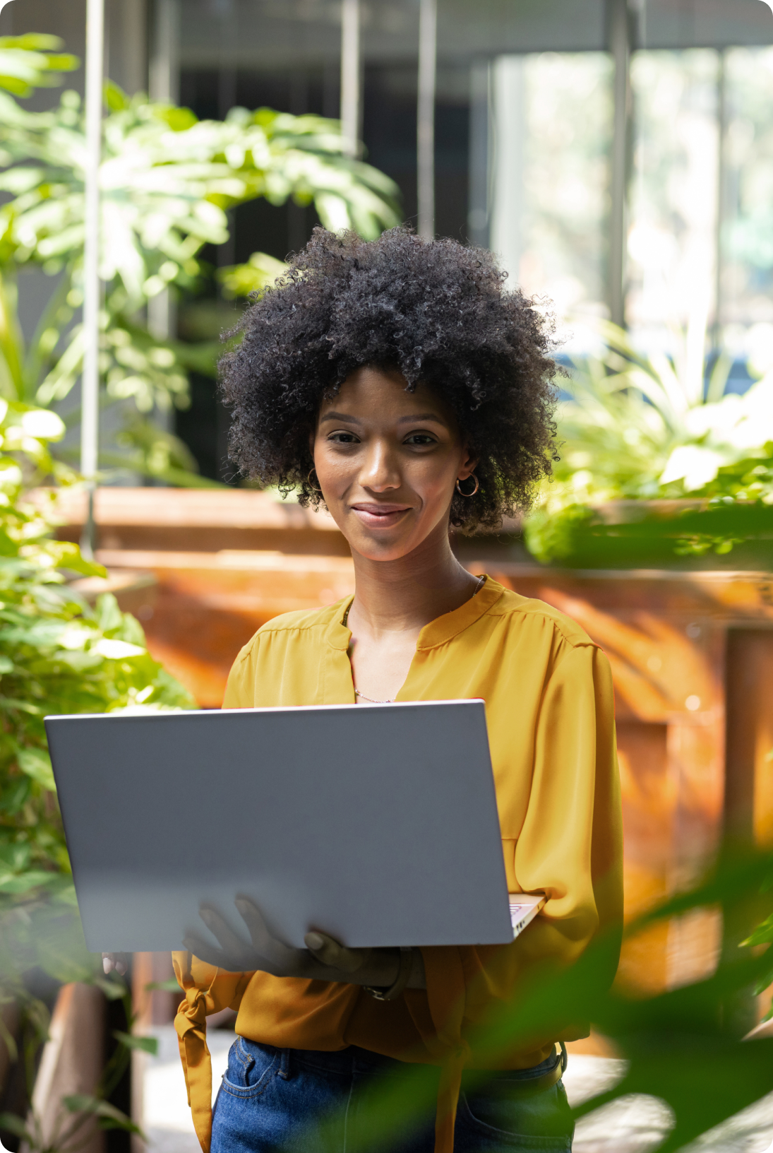 Mulher segura notebook em ambiente com plantas, usando blusa amarela e sorrindo levemente.