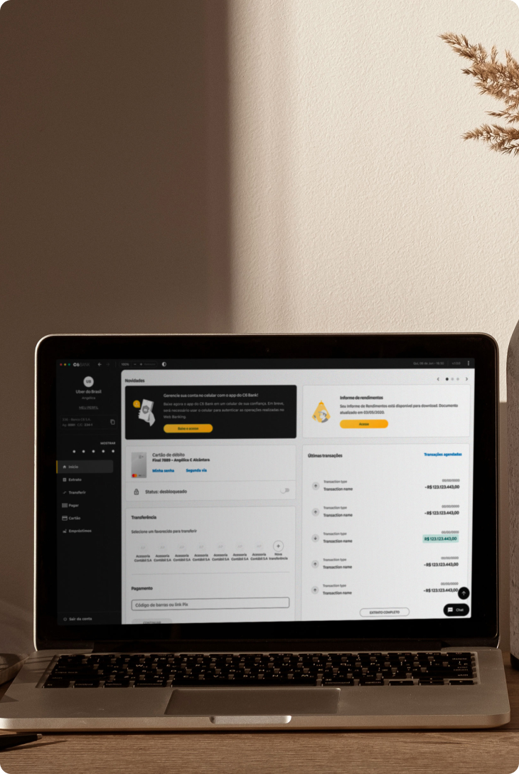 Notebook em mesa com tela do Web Banking aberta, exibindo informações de cartão e transações recentes. Ao lado, óculos, livros e vaso decorativo.