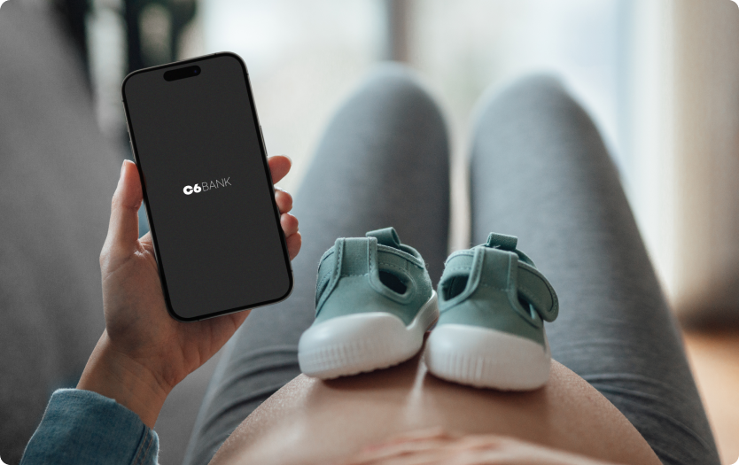 Mulher grávida segura um celular C6 Bank com sapatinhos sobre a barriga.