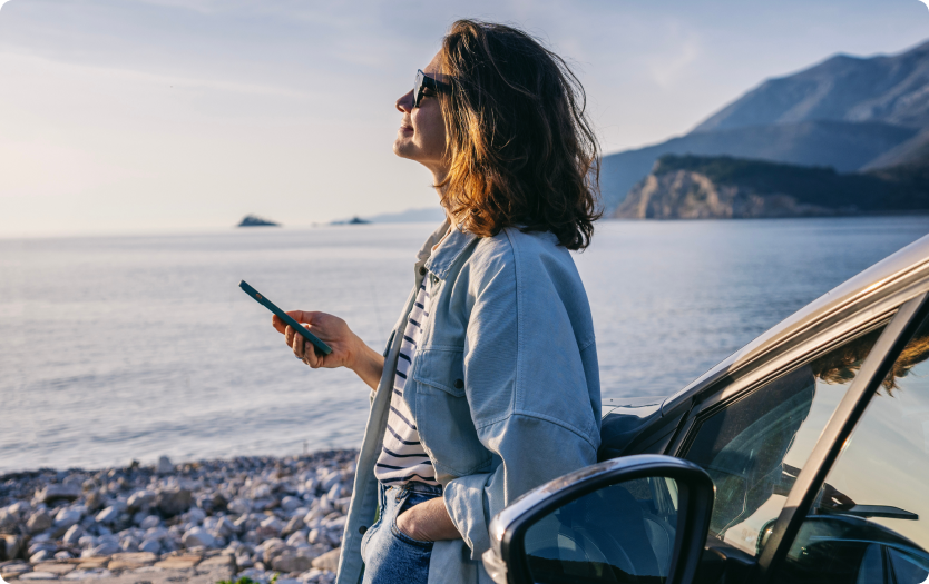 Mulher ao lado do carro segura celular e observa o mar com montanhas ao fundo.