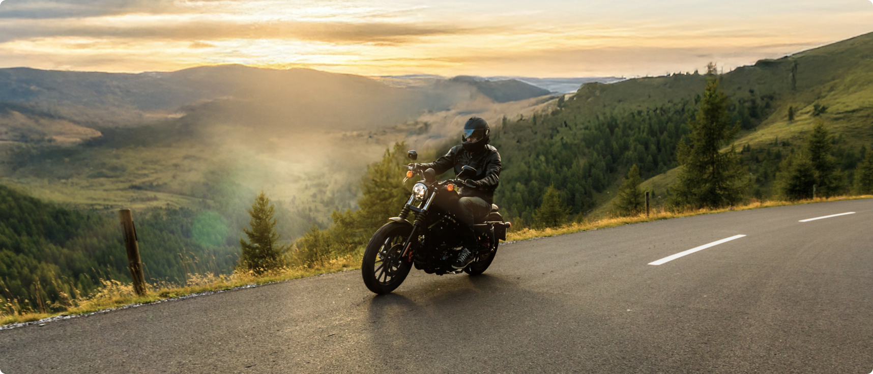 Motociclista em estrada de montanha ao entardecer, com paisagem de vales e colinas ao fundo.