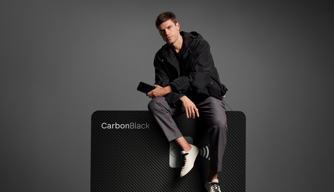 Homem sentado sobre cartão C6 Carbon ampliado, em fundo escuro. Visual elegante e minimalista.