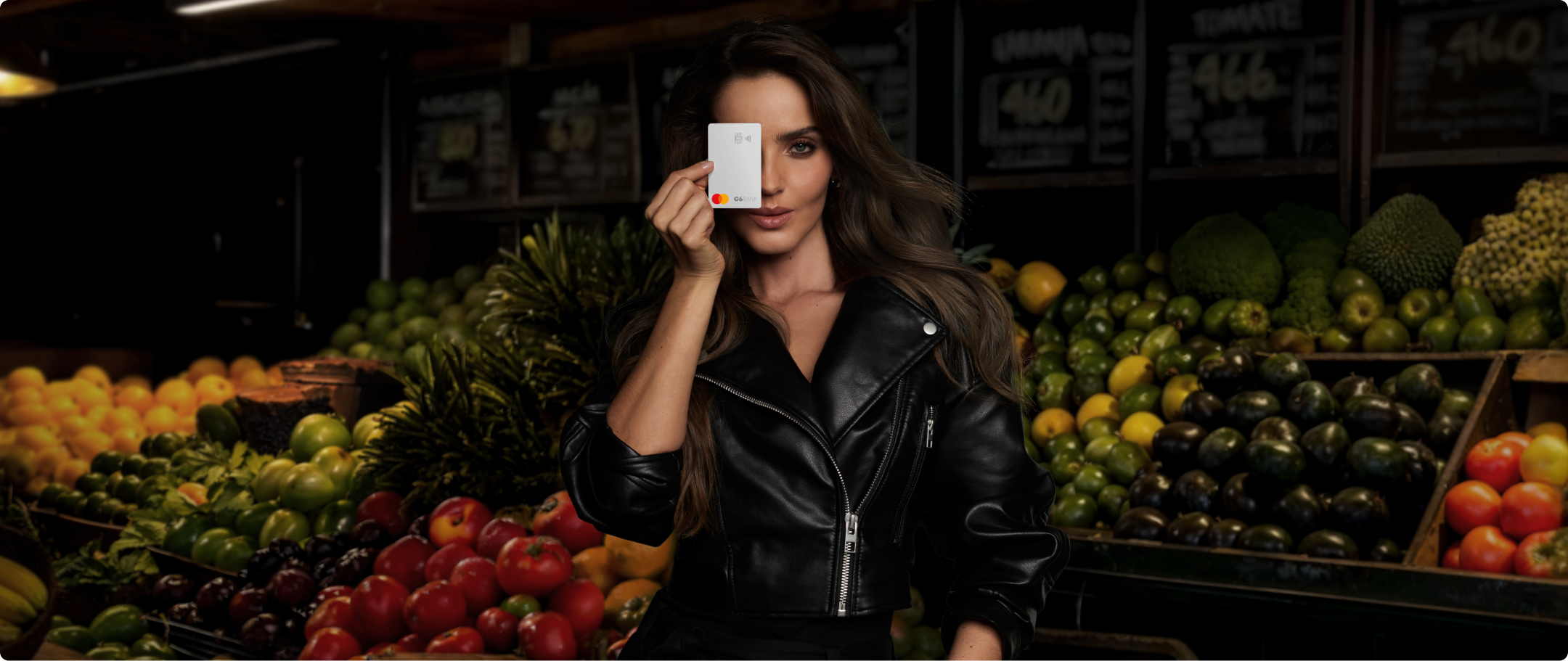 Mulher cobre um olho com Cartão C6 na cor Silver em feira iluminada, cercada por frutas. Cena com forte contraste.