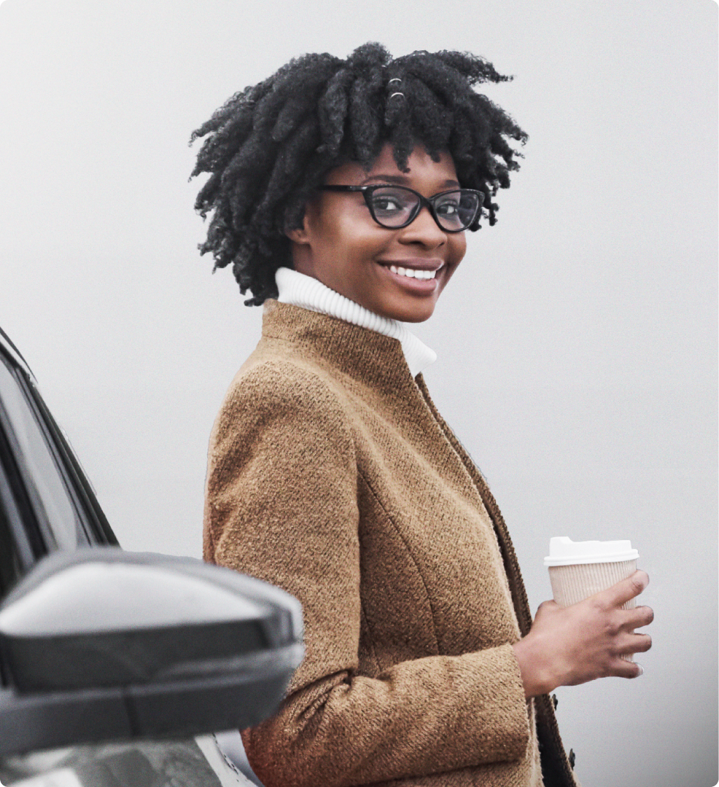 Mulher sorridente com café na mão, vestindo casaco e óculos, ao lado de um carro.