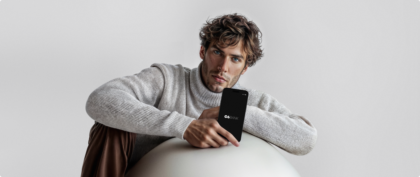 Fotografia de homem branco com cabelo de comprimento médio, ondulado e castanho, vestindo uma blusa de tricô branca e calça marrom. Ele está apoiado em uma esfera branca e segura um celular com a tela preta e o logo do C6 Bank no centro
