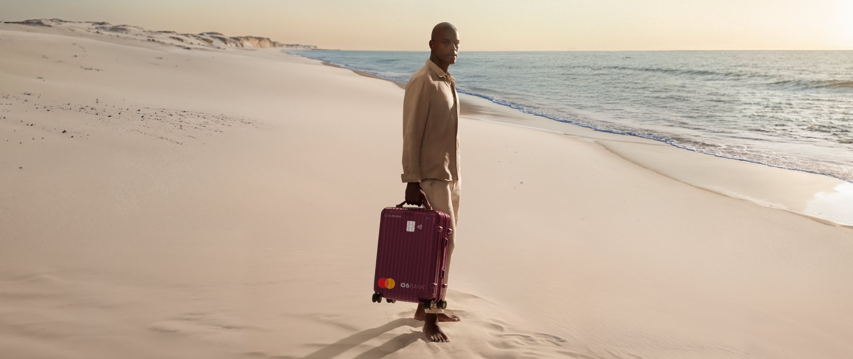 Homem na praia puxa mala com layout do C6 Conta Global. Cena ao entardecer transmite mobilidade e tranquilidade.