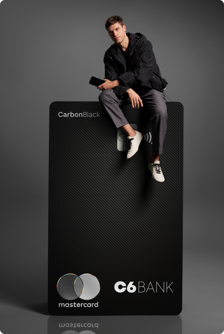 Homem sentado sobre cartão C6 Carbon ampliado, em fundo escuro. Visual elegante e minimalista.